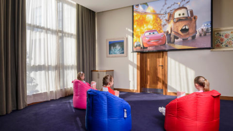 Children enjoy a cosy movie experience in a hotel’s bright media room with bean bags and a large screen showing an animated film.