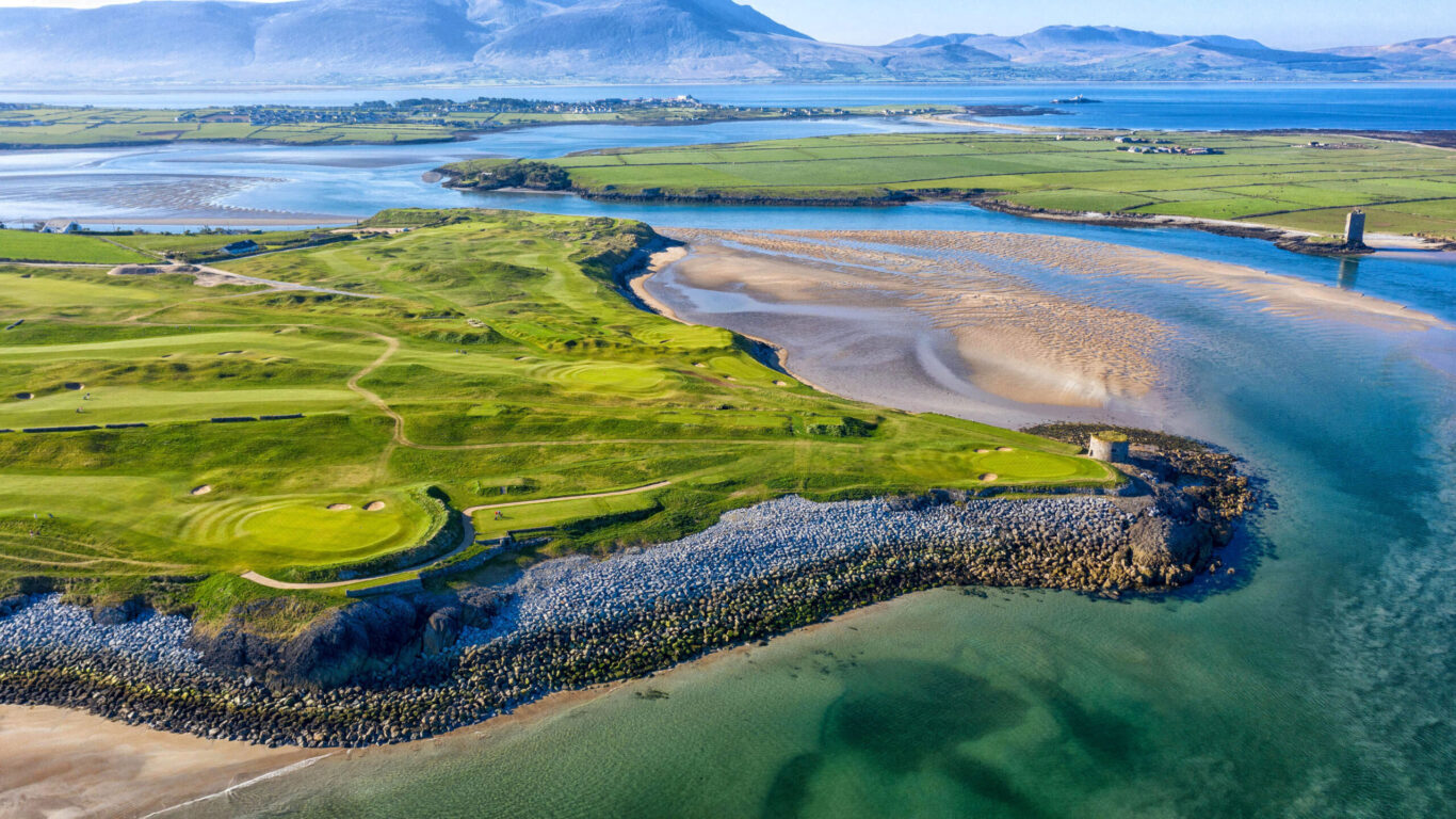 Aerial view of a coastal golf course with green fairways, sandy beaches, rock barriers, and surrounding water, with mountains in the distance under a clear sky.