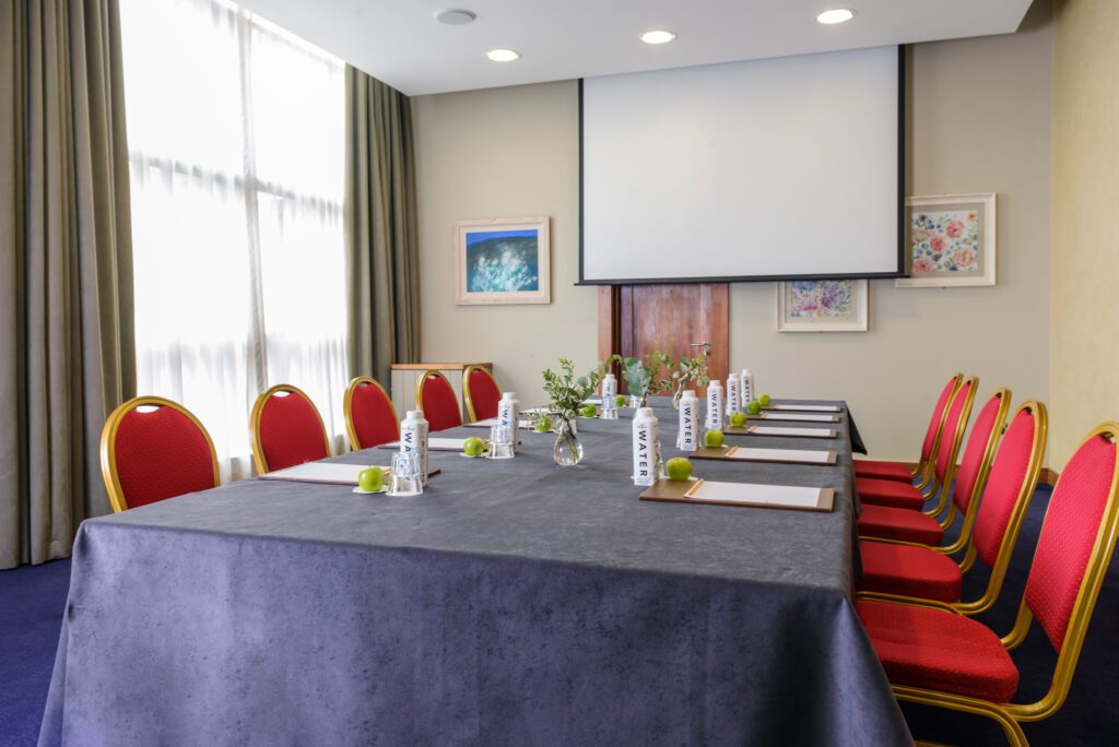 A conference room with a table set for a meeting, featuring red chairs, water bottles, notepads, green apples, and a projection screen at the front.
